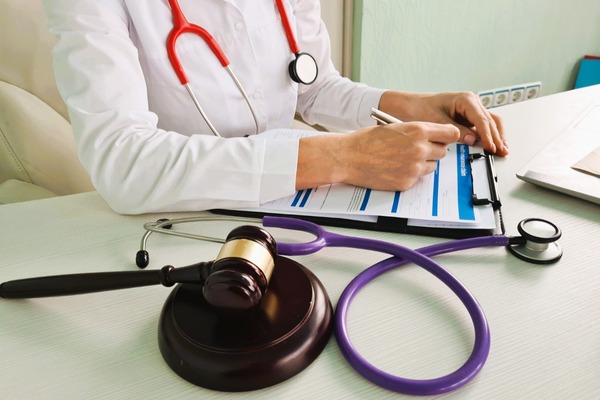 Médico con bata escribe en un formulario; junto a él, un martillo judicial y un estetoscopio. Relación médico-legal.