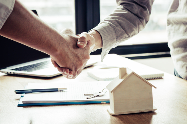 Dos personas se dan la mano frente a un contrato con llaves y maqueta de casa, ilustrando herencia de vivienda tras fallecimiento del cónyuge.