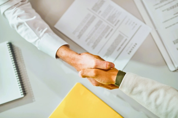 Dos personas se dan la mano sobre documentos legales, representando la firma de un pacto de socios en un acuerdo empresarial.