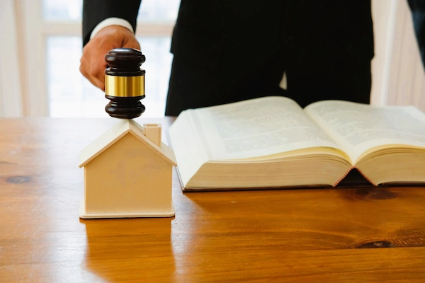 Persona con mazo judicial junto a una maqueta de casa y un libro abierto, simbolizando la subasta de inmueble en copropiedad.
