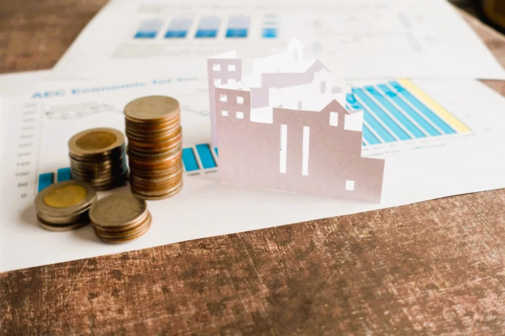 Monedas y viviendas sobre gráficos financieros que representan la plusvalía municipal en Madrid tras la venta o herencia de un inmueble.